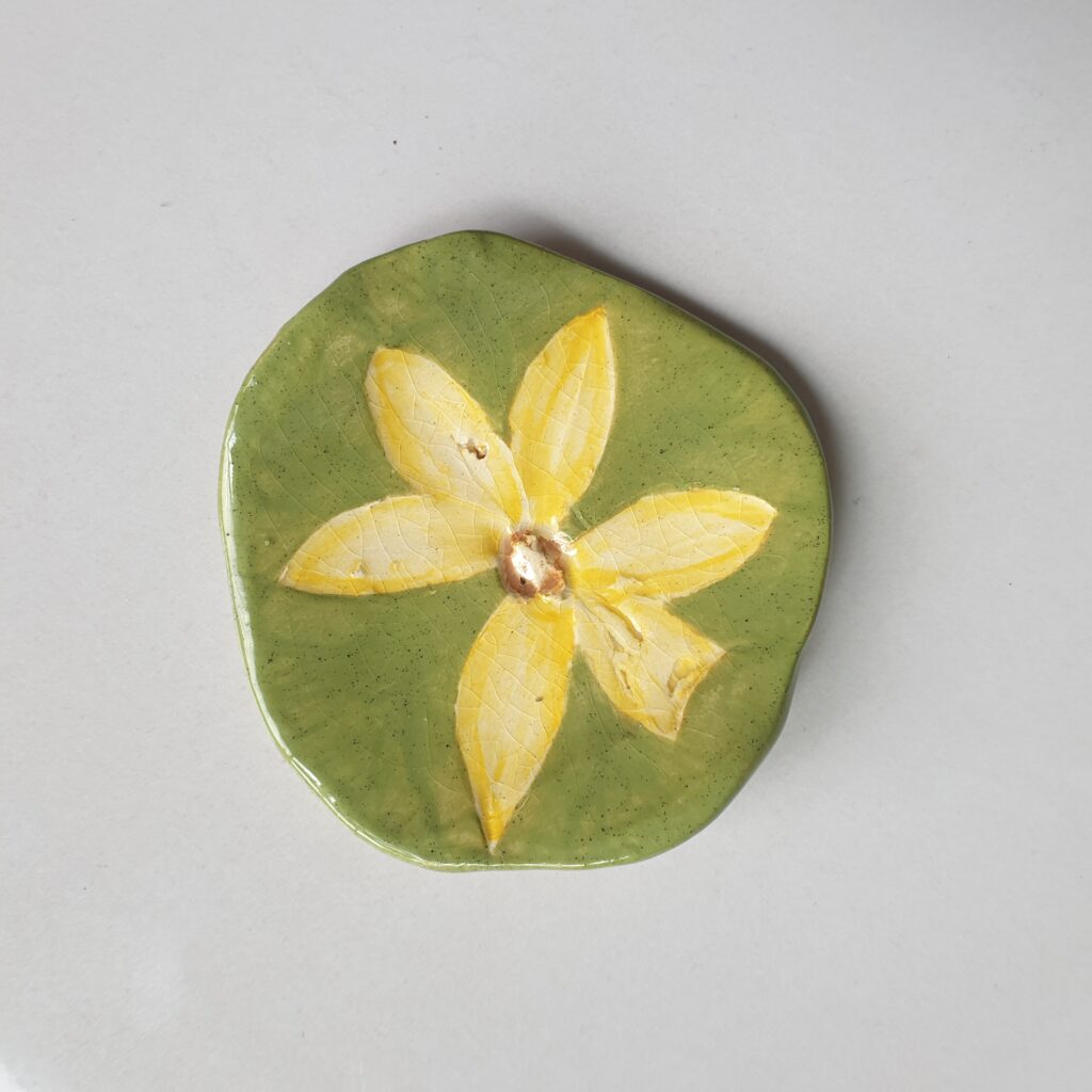A ceramic plate featuring a painted yellow flower on a green background using an imprint from nature.