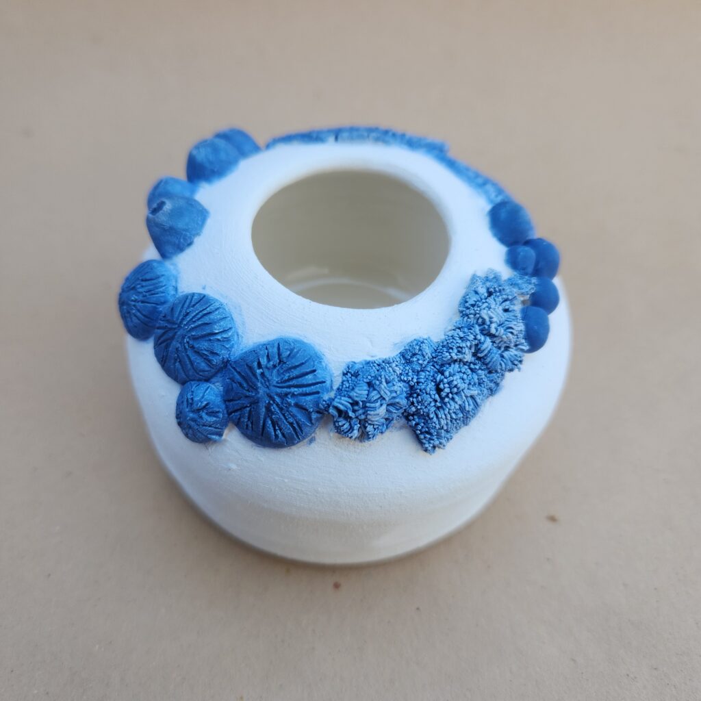 A small, round ceramic wheel thrown vessel with textured blue embellishments on a white surface.